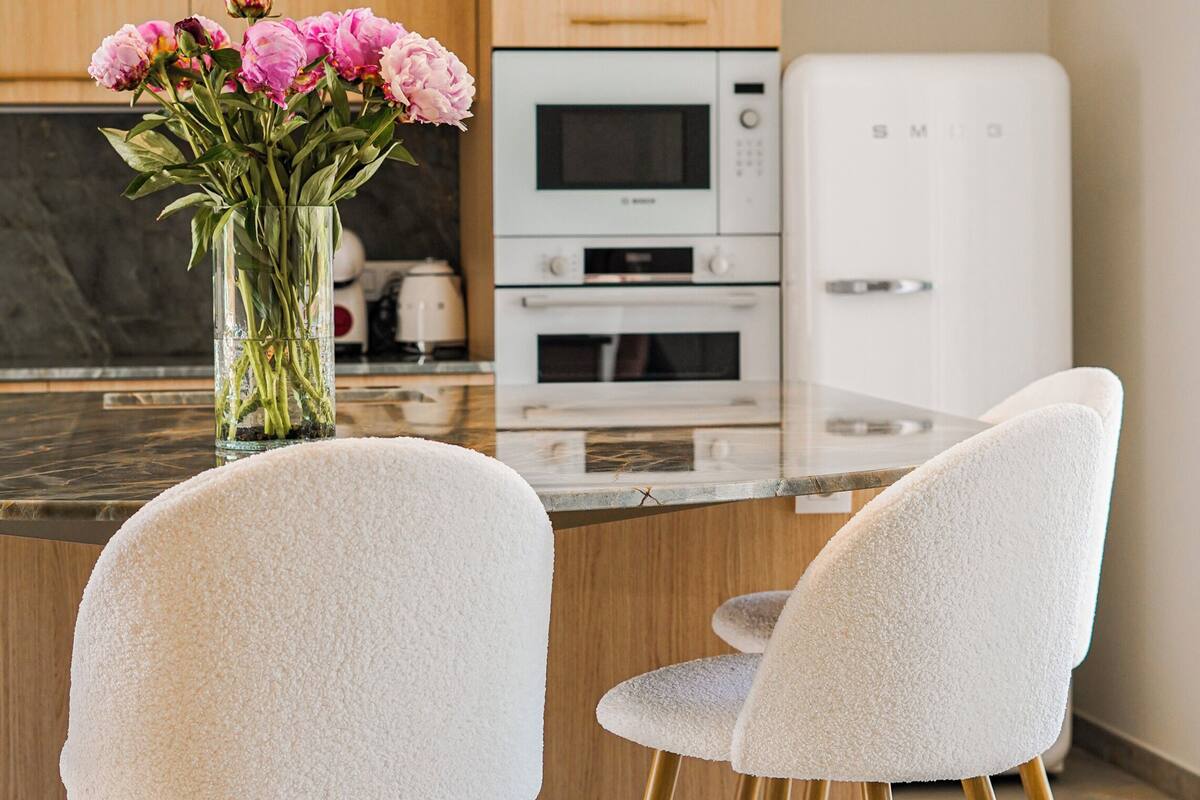 Een moderne keuken met een centraal eiland, twee barkrukken, een vaas met bloemen en inbouwapparatuur.