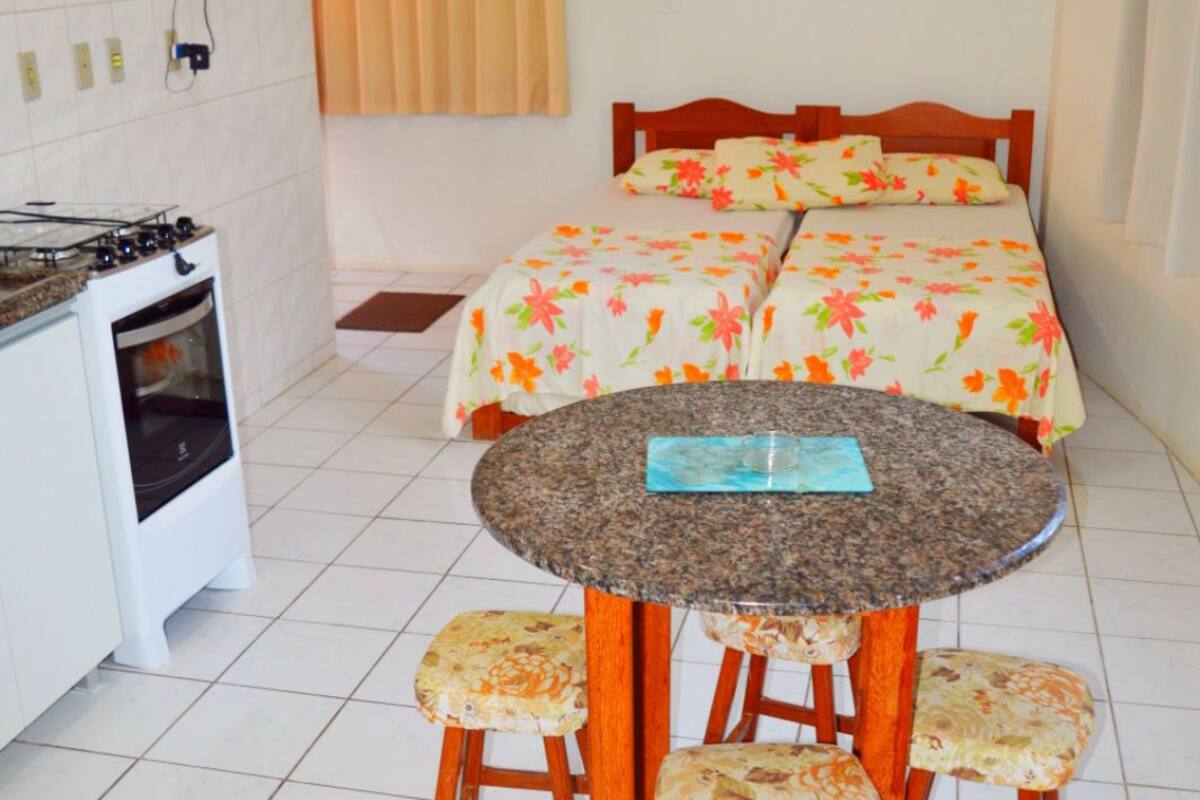 Une pièce comprenant un lit, une table à manger avec quatre chaises, un petit coin cuisine avec une cuisinière et un ventilateur de plafond.