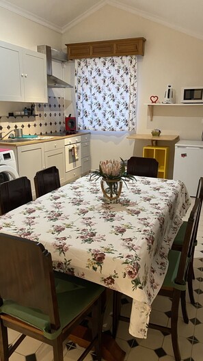 A dining area with a table set for four, surrounded by chairs. A floral tablecloth covers the table, and a vase with flowers is placed in the center. The room has a kitchen area with cabinets, a microwave, and a refrigerator.