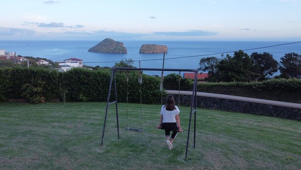 Eine grüne Außenanlage mit Schaukeln, einer Steinmauer und Blick auf das Meer mit zwei Inseln.
