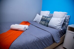 A neatly made bed with a grey and orange blanket, white and patterned pillows, and a white cabinet to the right. - Annex Lodge (Cape Town)
