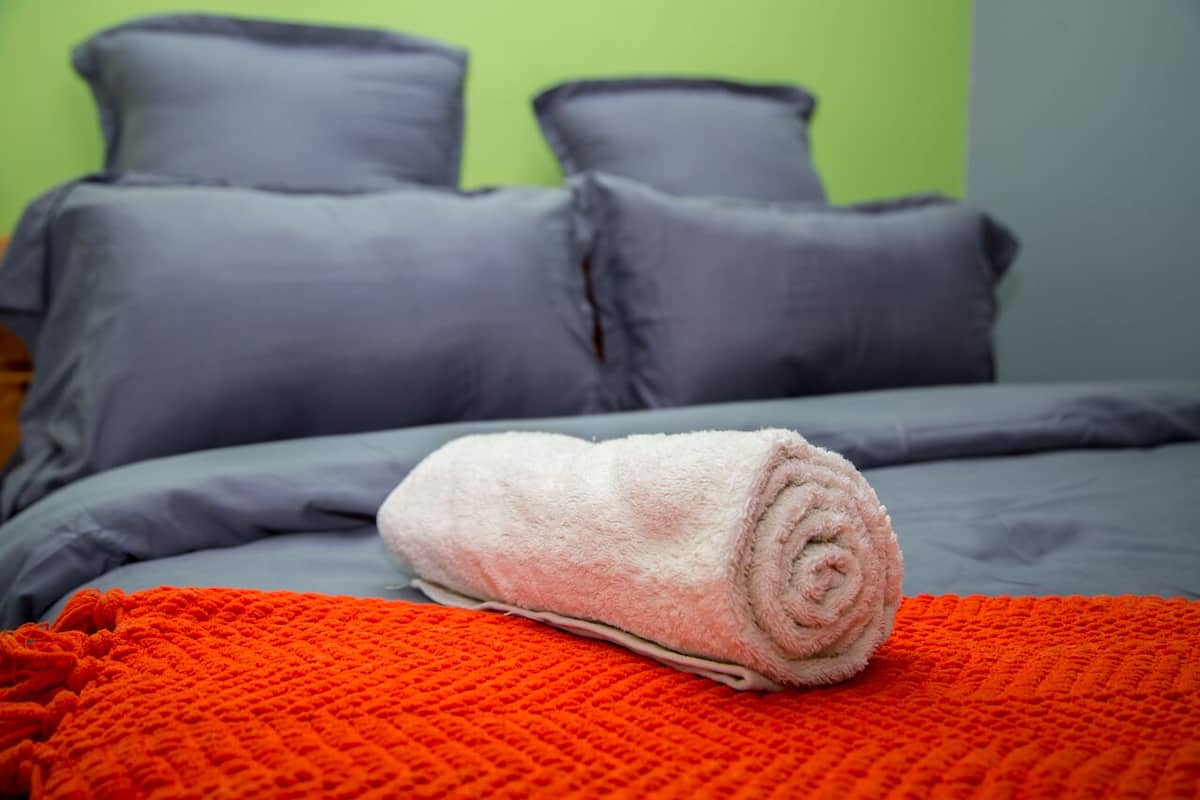 Una cama con ropa de cama gris, una toalla enrollada y una manta naranja.