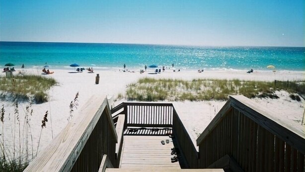 A beach with a wooden boardwalk leading to the shore, clear skies, and calm sea.