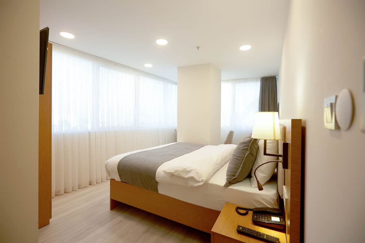 A modern hotel room with a wooden bed, a bedside table with a phone and remote, and a large window with sheer curtains.