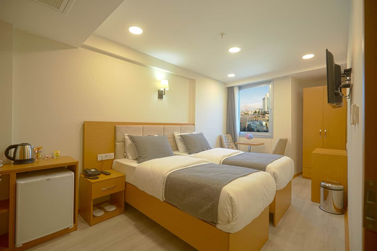 A modern hotel room with two beds, a small desk, a refrigerator, a TV, and a window with a city view.