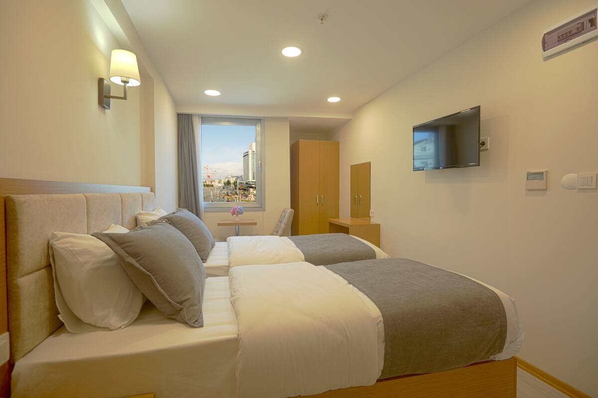 A hotel room with two beds, a TV, a window with a view of a cityscape, and a bathroom with a toilet and sink.