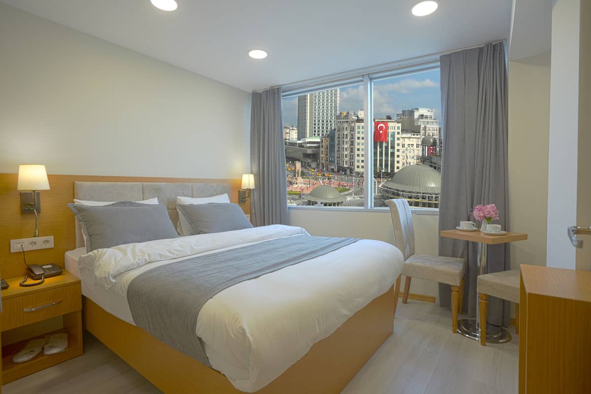 A modern hotel room with a large bed, bedside tables, a desk, and a view of the cityscape.