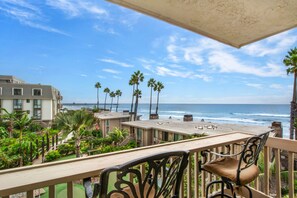 A balcony with a view of the ocean, palm trees, and a building with a beach in the background. - Front Row Ocean View. Studio, Luxury Remodel. E210 North Coast Village (Oceanside)