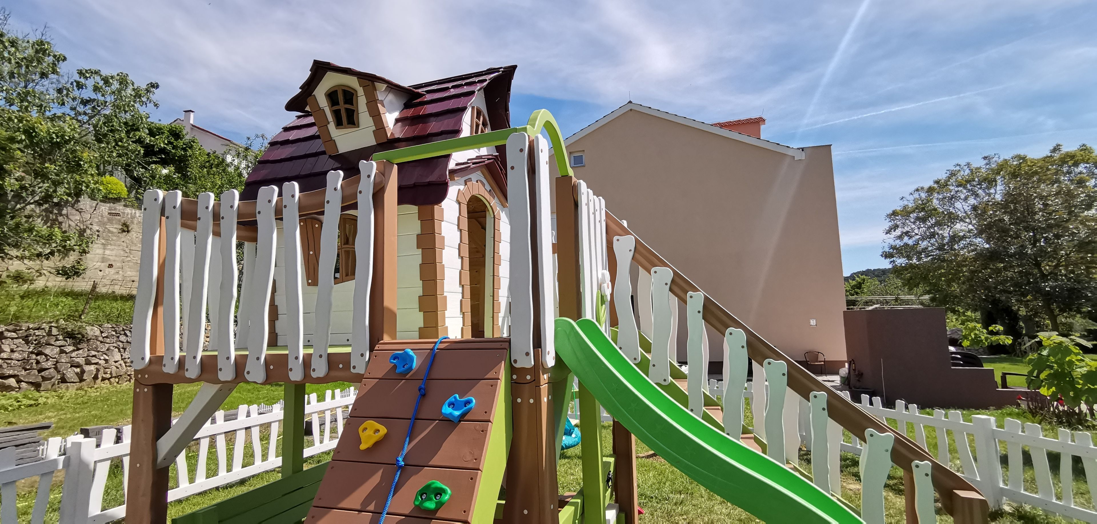 A children's play structure with a slide and climbing wall, surrounded by a white picket fence.