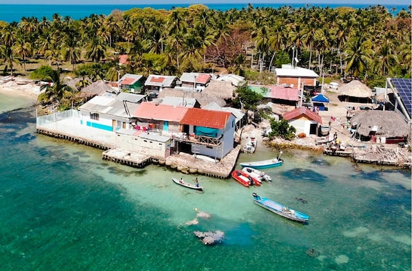 An aerial view of a coastal village with thatched-roof houses, a pier, and clear turquoise waters.