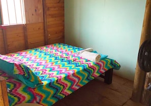 A small, single-bed room with a colorful zigzag-patterned bedspread, a wooden bench, and a standing fan.