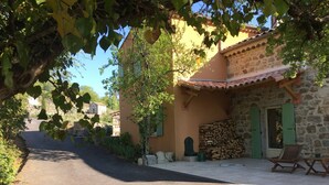 Un chemin étroit mène à un bâtiment en pierre surmonté d’un toit de tuiles, avec une pile de bois de chauffage et une chaise en bois placée à l’extérieur.