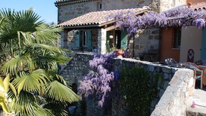 Un bâtiment en pierre avec un toit en tuiles, un petit balcon orné de fleurs violettes et un jardinière blanche posée au sol.