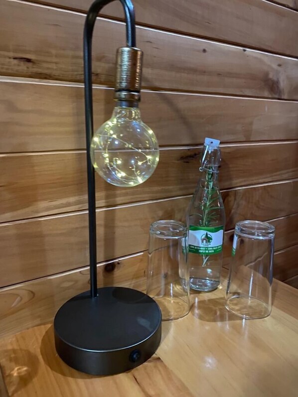 A modern lamp with a spherical glass shade, a black base, and a bottle of green liquid next to four clear glasses on a wooden surface. - Clusia Lodge (Copey)