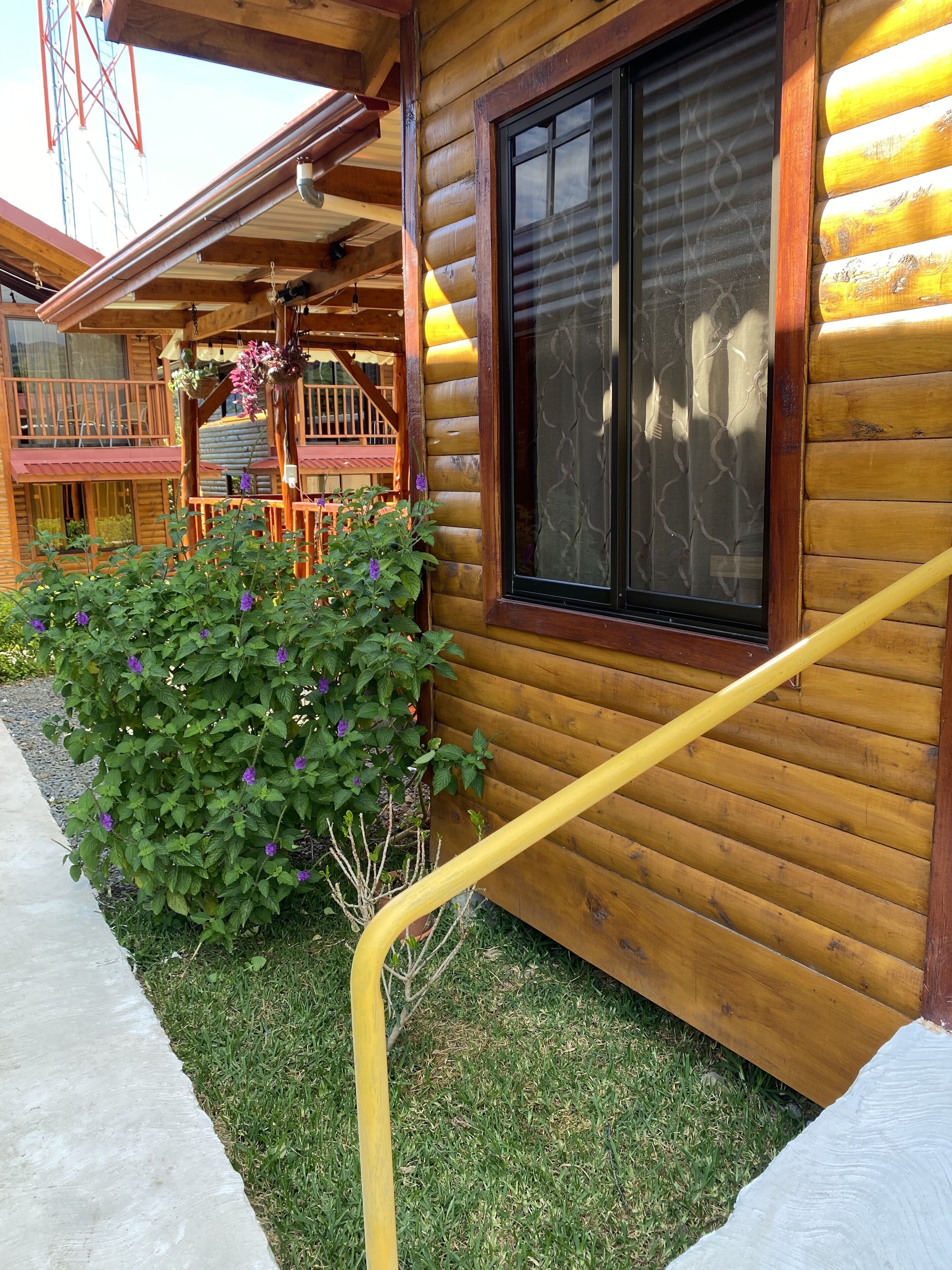 A wooden cabin with a balcony, a window with a patterned blind, and a yellow handrail.