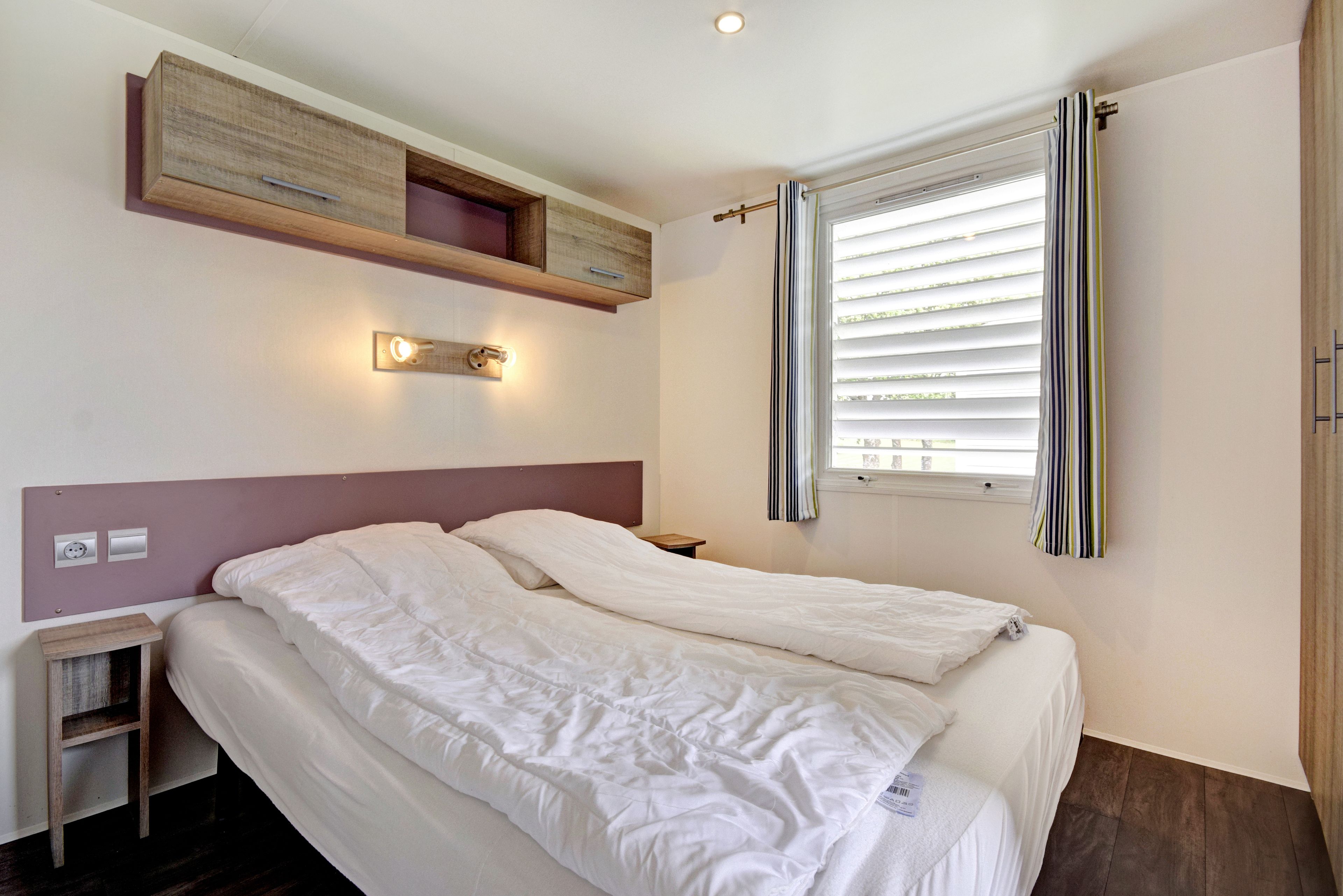 A double bed with a headboard, a bedside table, a wall-mounted shelf, and a window with blinds.