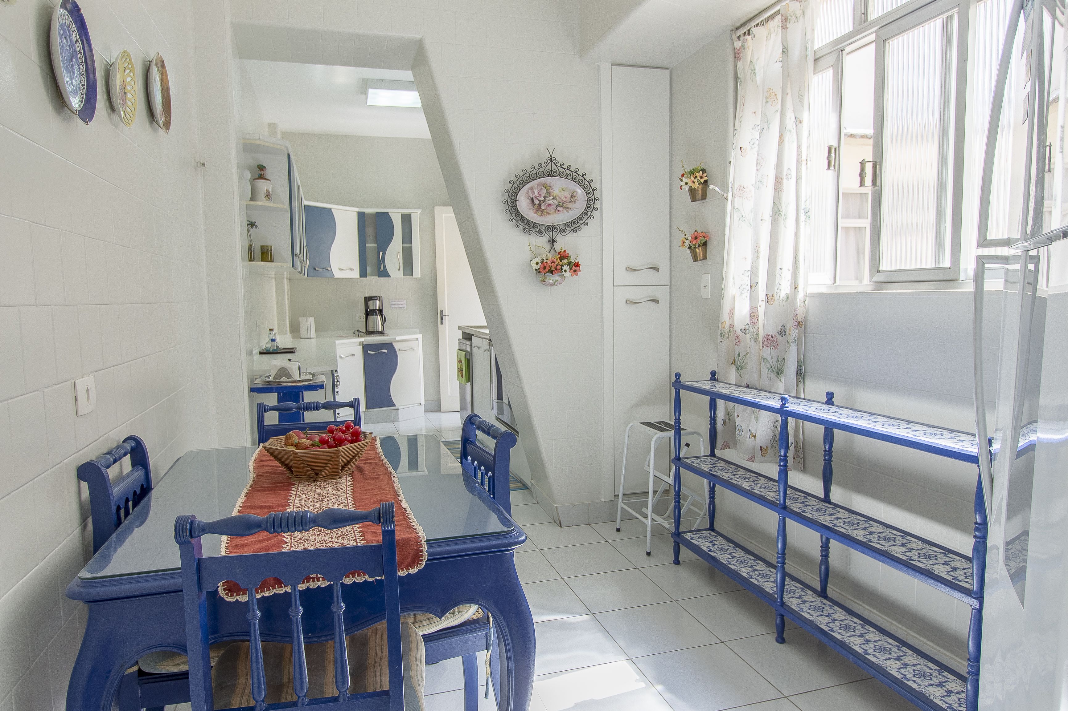 A dining area with a blue table and chairs, a white tiled floor, a kitchen in the background, and a window with a curtain.