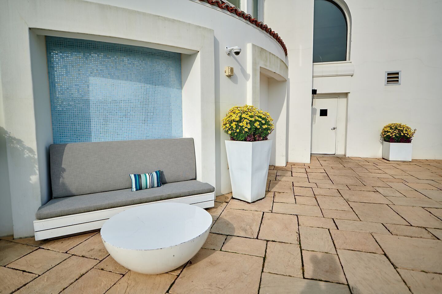 A modern outdoor seating area with a gray sofa, a white round table, and two potted plants with yellow flowers.