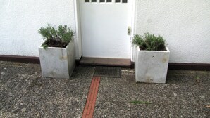 Une porte blanche, précédée d’un petit marchepied, et de part et d’autre, deux jardinières carrées ornées de plantes vertes.
