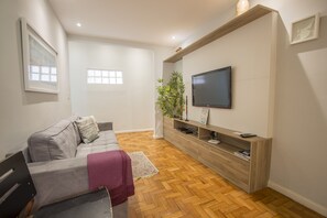 Sala de estar com sofá cinza, televisão sobre suporte de madeira e piso de madeira em padrão herringbone.
