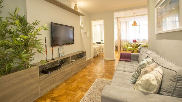 A modern living room with a grey sofa, wooden flooring, a TV on a wooden shelf, and a dining area with yellow chairs. - LineRio Praia de Copacabana 370.374 (Rio de Janeiro)
