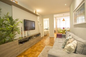 A modern living room with a grey sofa, wooden flooring, a TV on a wooden shelf, and a dining area with yellow chairs. - LineRio Praia de Copacabana 370.374 (Rio de Janeiro)