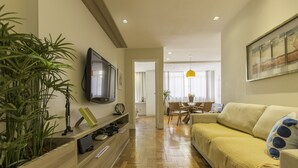 A modern living room with a wooden floor, a beige sofa, a TV mounted on the wall, and a dining area in the background. - LineRio Praia de Copacabana 370.374 (Rio de Janeiro)
