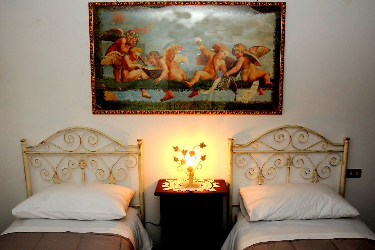 Un dormitorio con dos camas, una mesita de noche con lámpara y un cuadro clásico en la pared.