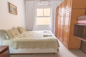 Een net opgemaakt bed met een sprei en kussens, een raam met gordijnen en houten kastjes.