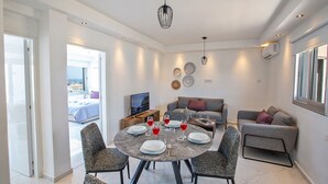 Un comedor moderno con una mesa redonda dispuesta para cuatro personas, rodeada de sillas tapizadas en gris. Se ve un sofá gris y una pequeña mesa de centro con un televisor al fondo.
