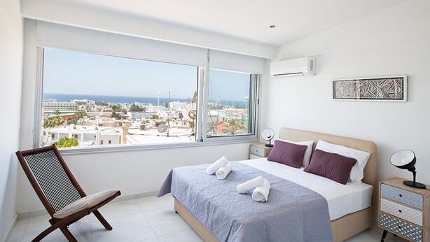 Un dormitorio con un ventanal grande, una cama con almohadas, una silla y una mesita de noche con una lámpara.