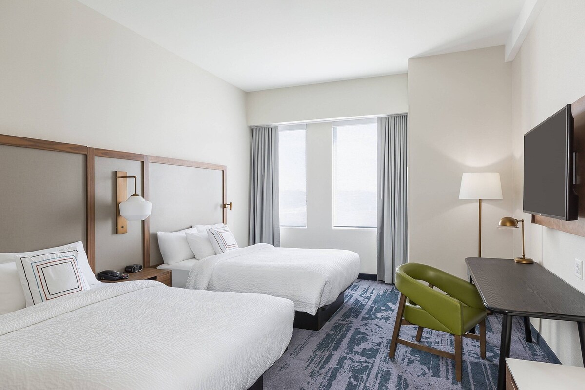 A hotel room with two beds, a desk, a TV, and a window with curtains.