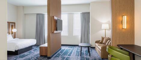 Habitación de hotel con cama, escritorio, una silla verde, sofá y televisor.