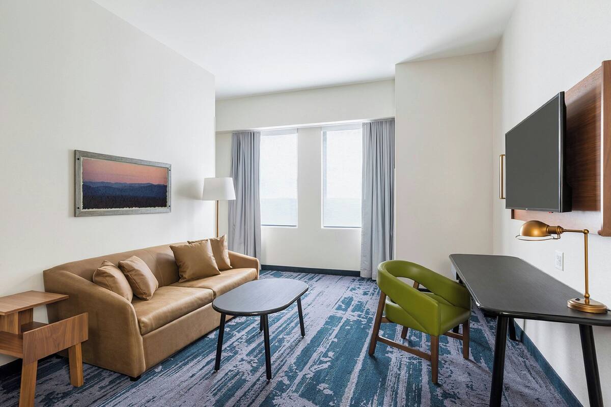 Une chambre d’hôtel avec un canapé, une petite table, un fauteuil vert, un bureau, une lampe, une télévision et une fenêtre avec des rideaux.