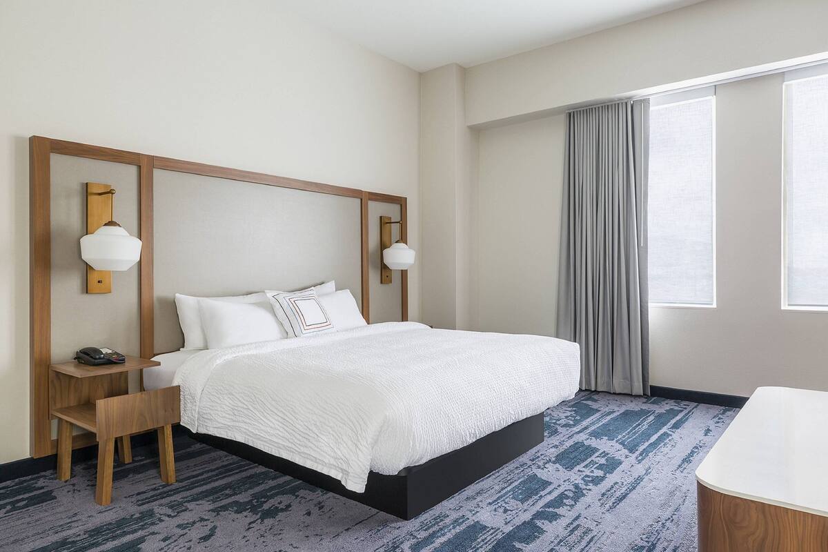 Une chambre d’hôtel avec un grand lit, une table de chevet en bois, un bureau et une chaise. Il y a une fenêtre avec des rideaux et une vue sur l’extérieur.