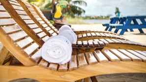 A wooden lounge chair with a rolled-up towel on it, a blue table, and a colorful bird-shaped chair in the background. - Luxury Beachfront Cabana with swim up pool bar (Placencia)