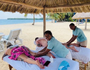 Un área para masajes en la playa con techo de paja, una camilla y una silla de descanso.