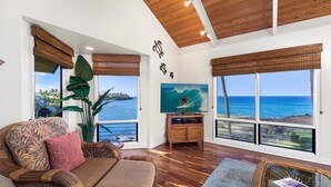 A spacious living room with a large window overlooking the ocean, a comfortable sofa, a television, and a large plant. - Kanaloa at Kona 3304: Delightful Oceanfront Condo w/ AC, Pool & Spa (Kailua Kona)