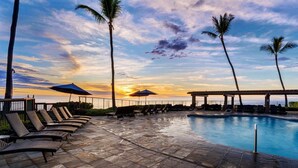 A poolside area with lounge chairs and umbrellas, a clear view of the sunset, and palm trees. - Kanaloa at Kona 3304: Delightful Oceanfront Condo w/ AC, Pool & Spa (Kailua Kona)