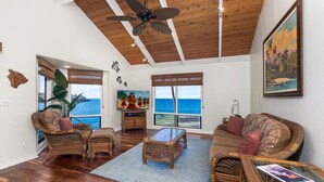 A spacious living room with a large window overlooking the ocean, featuring wicker furniture and a ceiling fan. - Kanaloa at Kona 3304: Delightful Oceanfront Condo w/ AC, Pool & Spa (Kailua Kona)