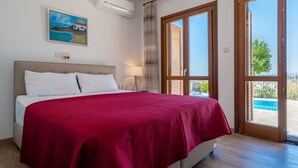 A bedroom with a large bed, a painting on the wall, an air conditioner, and a view of the outside. - Stunning 3 bedroom villa 'BZ01' with private pool, stunning views, communal pool and resort facilities, Zephyros Village on Aphrodite Hills Re (Kouklia)