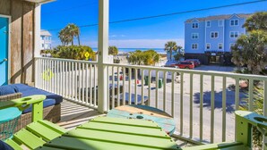 A balcony with green chairs and a wicker sofa overlooking a beach and a building with multiple windows.