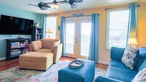 A living room with a beige armchair, a blue sofa, a small ottoman, a flat-screen TV, and a bookshelf.