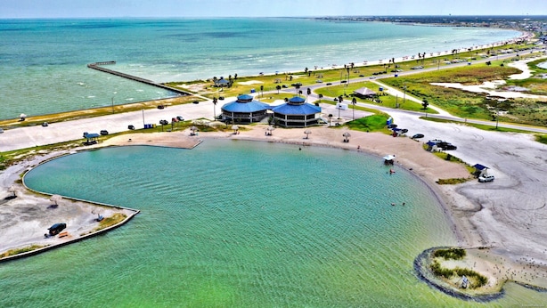 Eine Küstenlandschaft mit einem großen Gewässer, einer Pier und einem Strand mit Sonnenschirmen und einigen Personen.