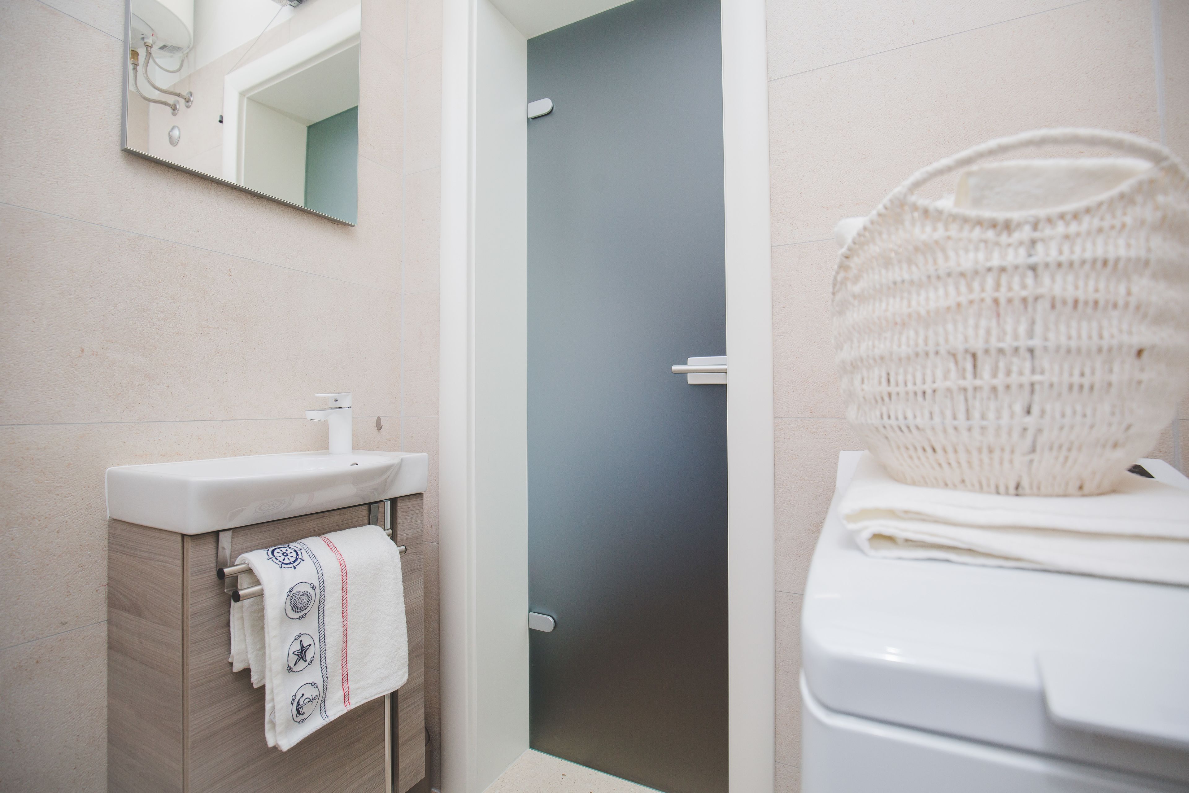 Une salle de bain avec un lavabo blanc, un porte-serviettes avec des serviettes, un miroir et un panier en osier posé sur les toilettes.