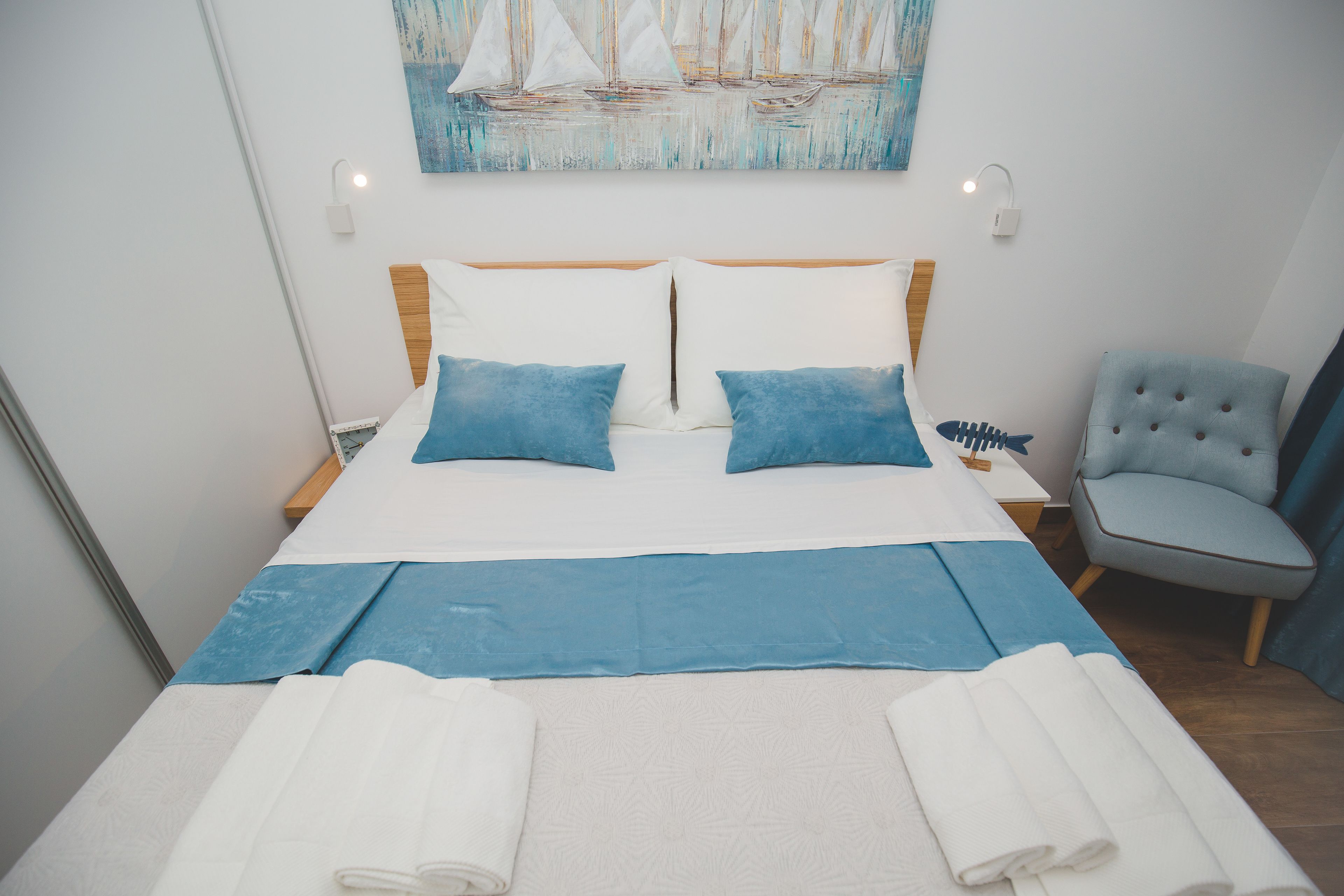 Uma cama bem arrumada com roupa de cama azul e branca, uma cadeira e um quadro de barcos à vela na parede.
