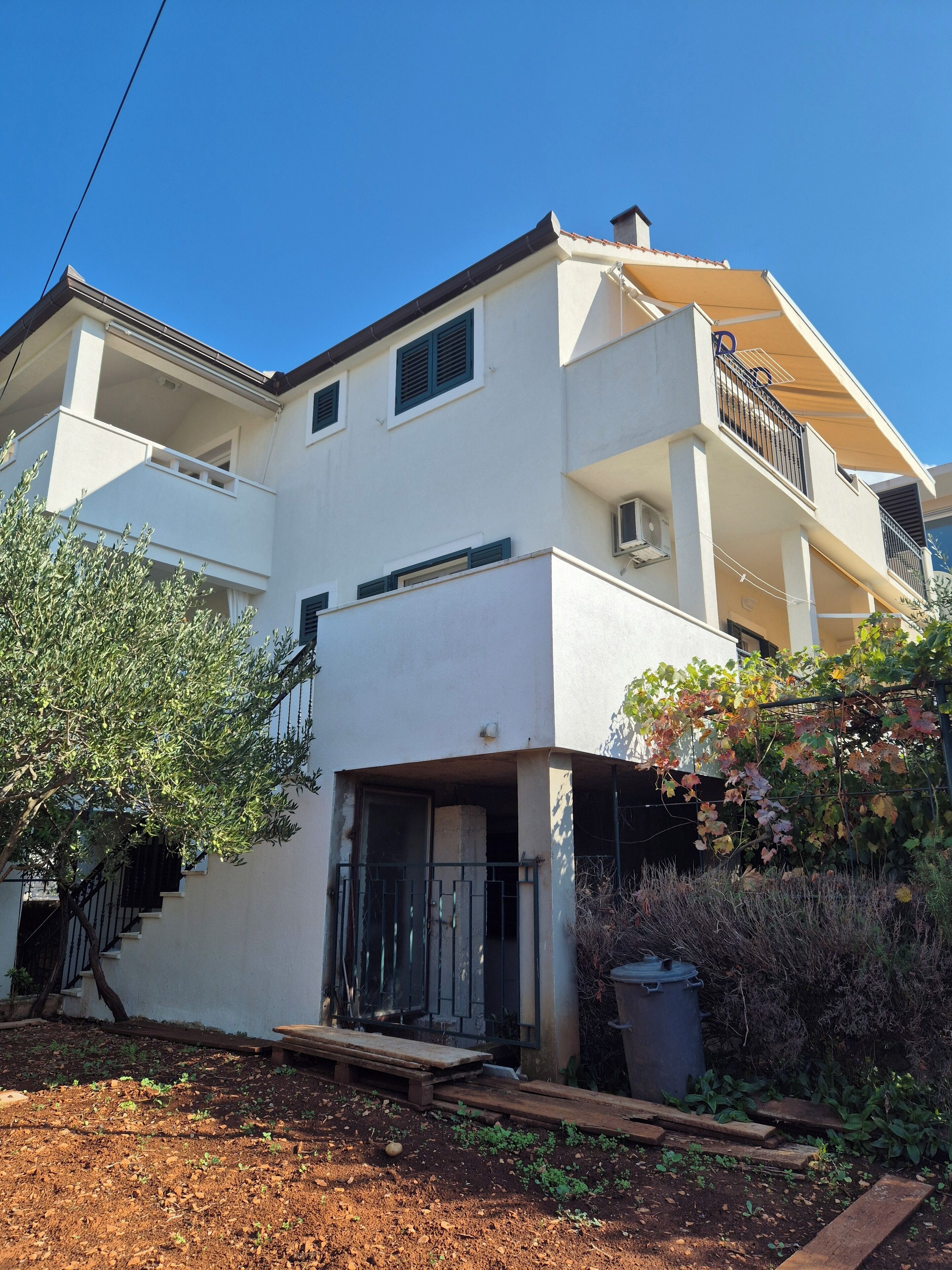 A two-story white building with a balcony, a staircase, and a garden area with plants and a trash bin.