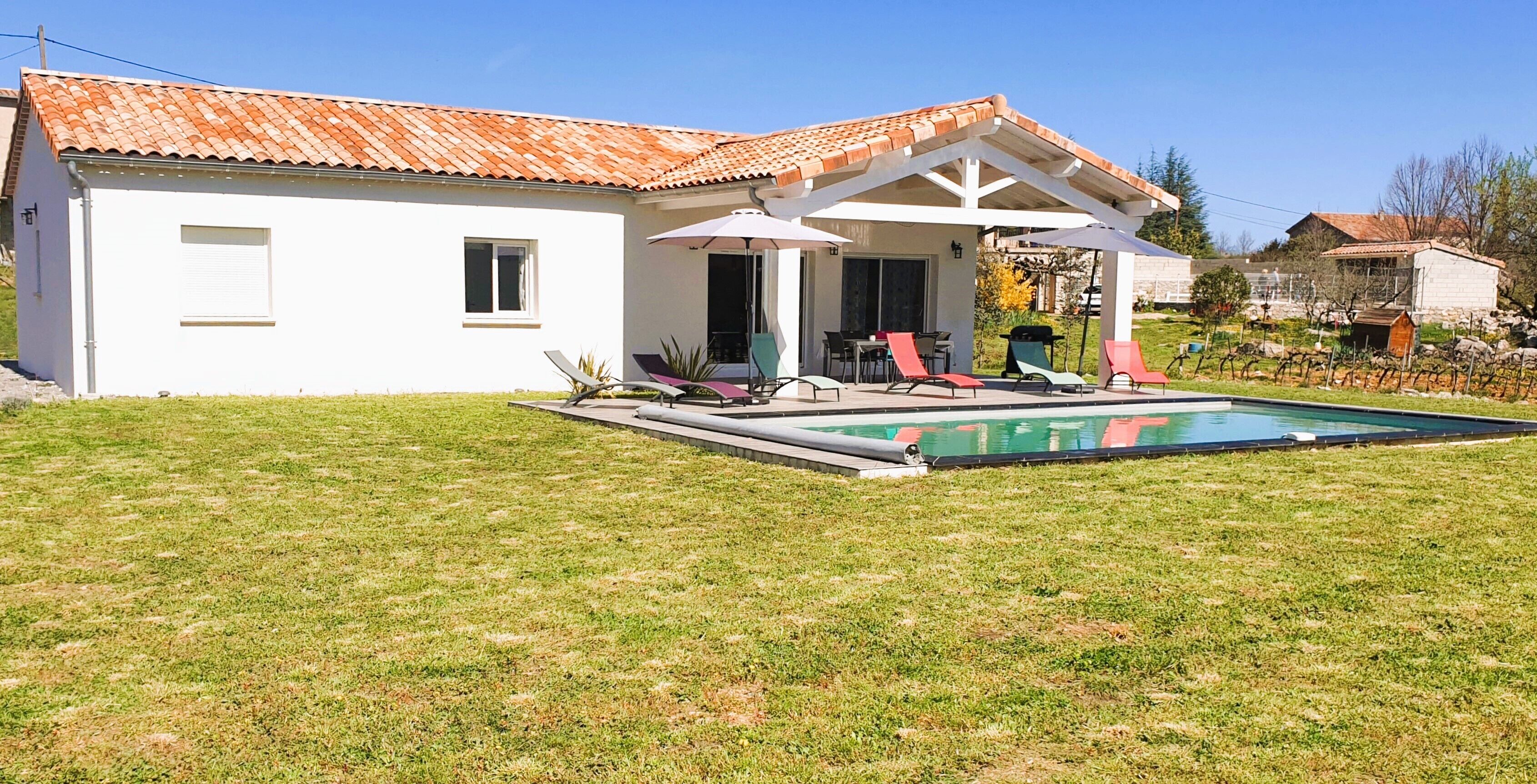 A white building with a tiled roof, a swimming pool, and a covered patio area with chairs.