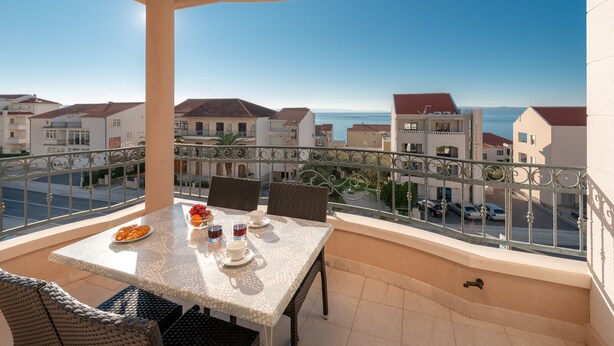 A balcony with a table set for two, overlooking a street and buildings. - Villa Tucepi Apartment 2 (4+1) (Tucepi)