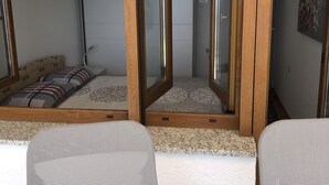 Ett litet rum med en säng, ett litet bord och en stol. Det finns en trädörr med glaspaneler och en väggmonterad luftkonditioneringsanläggning.